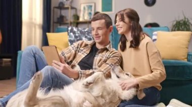 Beyaz ırktan insanlar evlerinde birlikte dinleniyorlar. Çekici koca tablet kullanırken güzel kadın yerde yatarken komik labrador 'u okşuyor. Aile fotoğrafı çeken sevimli bir çift..