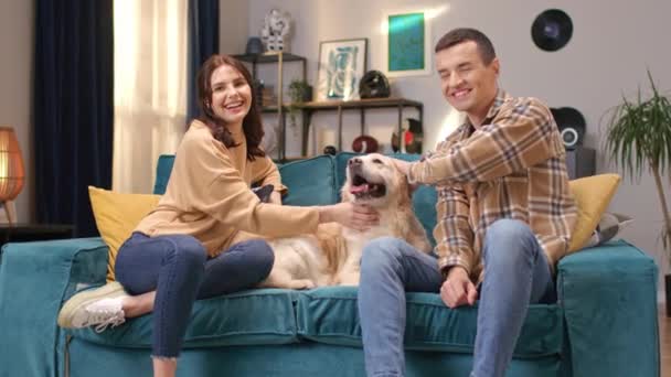 快乐的白种人夫妇与他们的毛茸茸的狗坐在沙发上的房间 人们兴高采烈地微笑着 爱抚着拉布拉多人 照顾他们的家畜 — 图库视频影像