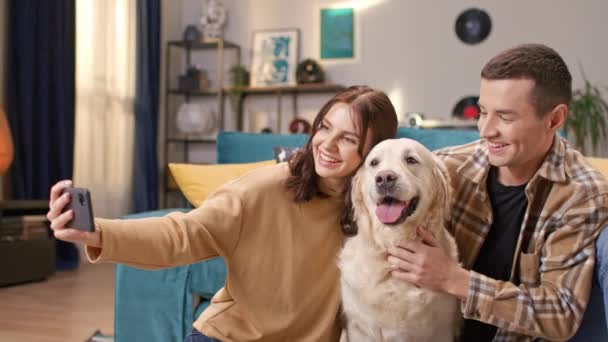 満足のいくカップルは 部屋で彼らの素敵な犬と家族の写真を撮ります 人々は床に座り 幸せな犬を抱きしめる 持っている 携帯電話 Petting ラブラドール — ストック動画