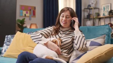 Köpeğiyle koltukta oturan neşeli bir kadın. Birisiyle telefonda iletişim kurmak. Evcil hayvanlara sarılıp okşamak. Şirin köpek sahibinin bacaklarının üzerinde yatıyor ve uyumaya hazır..