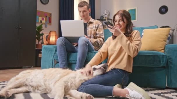 年轻的白人男子在笔记本电脑上工作 而他的积极妻子则在手机上给他们的爱玩的狗拍照 金色的拉布拉多 舌头放在主人的腿上 使用技术设备的人 — 图库视频影像