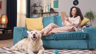 Güzel görünümlü beyaz kadın sahibi ve sevimli köpeği oturma odasında dinleniyor. Güzel kadın tablet aygıtı ve web siteleri kullanırken komik labradoru yerde yatıyordu..