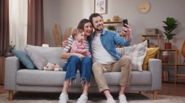 Gülümseyen aile oturma odasında kanepede dinleniyor. Genç evli çift küçük kızıyla eğleniyor. Baba, karısı ve çocuğuyla selfie çekiyor. Küçük çocuk telefon kamerasına poz veriyor. Mutlu aile.