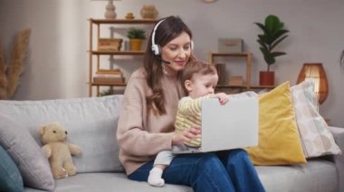 Küçük kız işe burnunu sokan anne. Mikrofonlu kulaklık takan bir kadın. Bebek bilgisayarı kapatıyor. Neşeli anne kameraya gülümsüyor. Annem çocuk elindeyken çalışmaya çalışıyor..