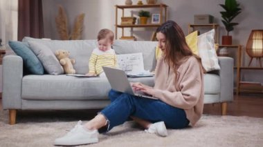 Halıda oturan ve dizüstü bilgisayarı olan bir kadın. Anne internette çalışıyor ve kızına bakıyor. Küçük kız kanepede oturuyor ve iş belgeleriyle oynuyor. Anne ve bebek evde..