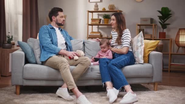 妻子和丈夫坐在沙发上讨论 年轻夫妇坐在沙发上聊天 已婚夫妇花时间与女婴在一起 家人周末在家里休息 小孩在玩电话 — 图库视频影像