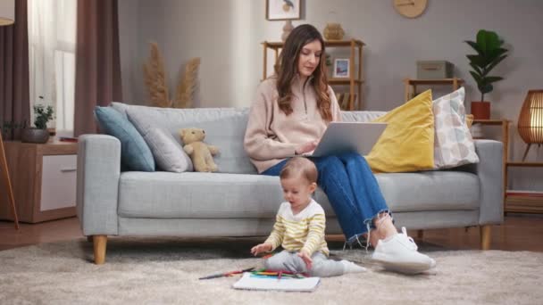 女人坐在沙发上拿着笔记本电脑 妈妈在网上工作照顾女儿小女孩坐在地毯地板上玩彩色铅笔 妈妈和小孩在家里 — 图库视频影像