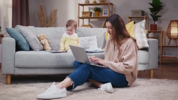 女人坐在地板地毯前 拿着笔记本电脑 妈妈在网上工作照顾女儿小女孩坐在沙发上玩商业文件 妈妈和小孩在家里 — 图库视频影像