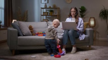 Evde çevrimiçi konferans veren bir kadın. Anne iş toplantısına katılıyor ve oğluyla ilgileniyor. Küçük çocuk renkli piramide yüzük takıyor. Mikrofonlu bir kadın..