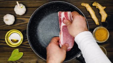 The cook puts the meat on the pan. Frying pan with meat steak, fresh garlic, olive oil, bay leaf, ginger, mustard peas, cumin on a table with a brown background with a wooden texture. Top view. Food