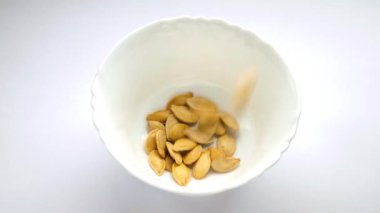 Balkabağı tohumları beyaz arka planda beyaz bir tabağa düşer. Ağır çekim. Kavrulmuş ve kurutulmuş bej kabak çekirdeği. Büyük sarı kabak çekirdekleri. Sağlıklı yiyecekler. Vejetaryen yemeği. Vejetaryenler için yemek. Yakın plan.