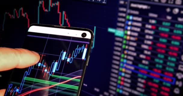 사업가는 컴퓨터 화면에 비즈니스 뉴스와 함께 자신의 스마트 폰에 금융 양초와 차트를 확인 — 비디오
