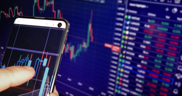 사업가는 컴퓨터 화면에 비즈니스 뉴스와 함께 자신의 스마트 폰에 금융 양초와 차트를 확인 — 비디오