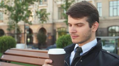 Keskin takım elbiseli genç bir adam ahşap bir bankta oturur, şık bir fincandan kahve yudumlarken dikkatlice uzaktan bakar. Bereketli yeşil ağaçlar ve hareketli bir şehirle huzurlu bir park.