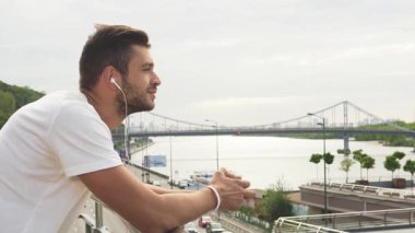 Koyu saçlı ve sakallı genç bir adam korkuluklara yaslanıyor, kulaklıklarından gelen melodilerde kayboluyor. Nehrin altında parladığı şehrin nefes kesen manzarasına bakıyor.