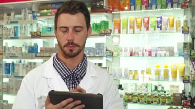 Gıcır gıcır bir tıbbi elbise giymiş odaklanmış bir erkek eczacı tabletindeki ilaç envanterini inceliyor. Etrafı düzgün düzenlenmiş renkli ilaç raflarıyla çevrili. Özenli bakışları ve profesyonel tavırları.