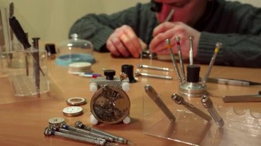 Odaklanmış orta yaşlı beyaz bir adam hassas aletler ve karmaşık saat parçaları ile çevrili hassas bir yineleyici saati ustalıkla tamir eder. Yakın plan saat yapma sanatını gözler önüne seriyor.