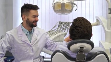 Genç bir beyaz dişçi, dişçi koltuğunda oturan erkek hastasına aynayı uzatırken sıcak bir şekilde gülümser ve dişçi tedavisinin sonuçlarını hevesle inceler. Parlak, modern klinik.