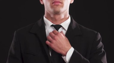 Zarif bir genç adam kravatını ince siyah bir arka plana karşı duyarlı, etkileyici ve sofistike bir şekilde ayarlar. Yarı görünür yüzü, geniş bir gülümseme ile tamamlanmış bir güven yayar.