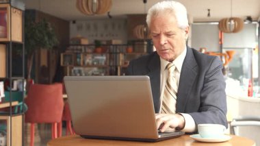Sivri takım elbiseli ve kravatlı yaşlı beyaz bir adam ahşap bir masada oturur ve kahvesini yudumlarken dizüstü bilgisayarını dikkatle analiz eder. Kafenin ılık havası, yumuşak ışıklandırma ve şık dekorla