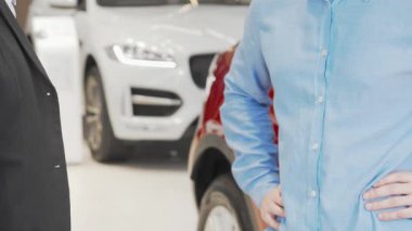 Memnun bir adam, başarılı bir araba alımını simgeleyen şık bir galeride iyi giyimli bir araba satıcısından araba anahtarı alır. Parlak, modern vitrinlerde parlak otomobiller sergileniyor.