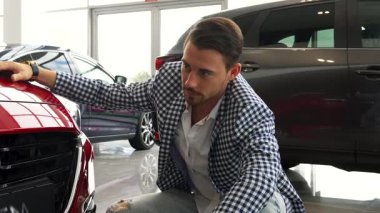 Koyu renk saçlı ve sakallı şık bir adam yeni arabasının yanında diz çöküp tamponunu tatmin olmuş bir gülümsemeyle teftiş ediyor. Şık bir ceket ve kot pantolon giymişti. Heyecan saçıyordu.