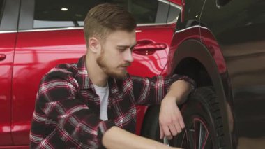 Şık sakallı neşeli genç bir adam parlak siyah bir arabanın yanında diz çöker, parlak tekerleklerini ve lastiklerini tatmin edici bir gülümsemeyle inceler. Baş parmak hareketi heyecan ve kendine güven yayıyor.