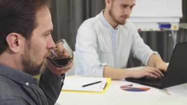 Yakın çekim, neşeli, olgun bir iş adamını kameraya gülümserken pozitiflik saçarak kahvesinin tadını çıkartırken yakalar. Bu sırada genç meslektaşı özenle arka planda çalışır. Çok canlı.