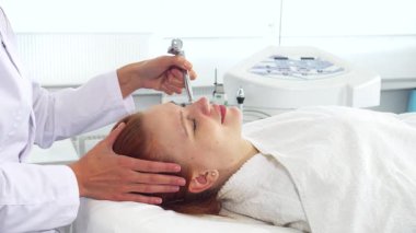 Yetenekli bir kozmetikçi, doğal güzelliğini arttırmak için sakin müşterilerinin yüzüne gelişmiş ekipmanları nazikçe uygular. Genç, çekici, beyaz kadın rahat bir şekilde rahat bir koltukta uzanıyor, gözleri...