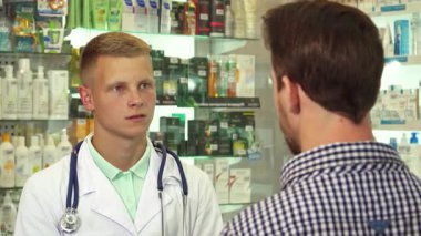 Parlak bir eczanede, beyaz önlüklü genç bir erkek doktor sıkıntılı bir erkek hastanın acı içinde başını sallamasını dikkatle dinliyor. Sahne, şefkatli bir sağlık hizmeti anını yakalıyor.