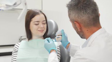 Neşeli bir kadın hasta modern bir diş sandalyesinde rahatça oturur olgun bir erkek dişçinin diş kalıbını göstermesi gibi gülümser. Bu ilgi çekici sahne diş bakımının önemini vurgular.