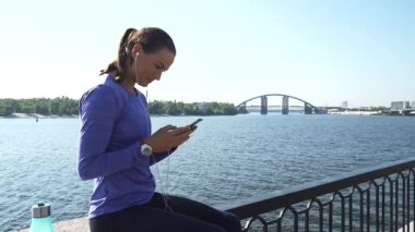 Uzun siyah saçlı, at kuyruklu neşeli genç bir kadın, canlı bir görüntülü sohbete dalarak nehir kenarındaki raylarda oturur. Canlı bir mor bluz ve parlak siyah taytlar giyip gülümsüyor.