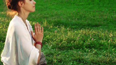 Güzel genç bir kadın yoga pozunda zarif bir şekilde oturur, ellerini nazikçe birbirine bastırarak meditasyon yapar. Etrafında yemyeşil çimenler ve yaz güneşinin yumuşak parıltısıyla, somutlaşır.