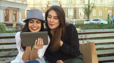 Biri şık gri şapkalı, diğeri dalgalı kahverengi saçlı iki modaya uygun bayan arkadaş, ahşap bir bankta otururken tablet bir ekranla büyüleniyorlar. Neşeli ifadeleri ve gündelik ifadeleri