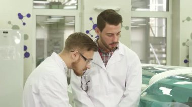 Parlak, modern bir laboratuvarda, iki genç erkek laboratuvar asistanı beyaz laboratuvar önlükleri ve lateks eldivenler giyerek iş akışları hakkında odaklanmış bir tartışmaya girişirler. Özenli ifadeleri...