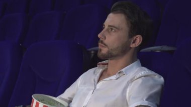 Loş bir sinema salonunda, koyu saçlı genç bir adam elinde patlamış mısırla ekranda oturuyor. Kameraya döner, parmağını dudaklarına koyar, filmin özünü somutlaştırır.