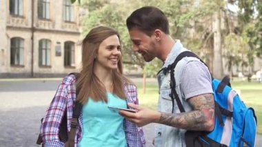 İki çekici Kafkasyalı öğrenci, neşeli bir genç kadın ve yakışıklı dövmeli bir adam, akıllı telefondan bir şey tartışırlarken keyifli bir anı paylaşıyorlar. Güneşli bir kampüs yolunda yan yana duruyoruz.
