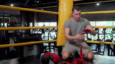 Kararlı genç bir Afrikalı erkek dövüşçü boks ringinin zemininde oturuyor, yumruklarını titizlikle kırmızı bandajlara sarıp yoğun bir antrenmana hazırlanıyor. Spor salonları enerjik atmosfer ve dövüşçülerin odaklandığı ifade ruhu somutlaştırıyor.