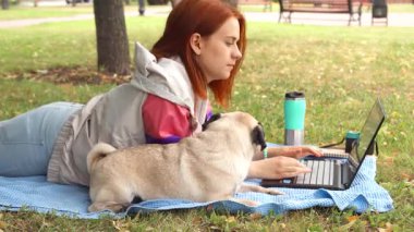 Canlı kızıl saçlı genç bir kız, güneş ışığı altındaki bir parkta mavi bir battaniyeye rahatça yayılmış, dizüstü bilgisayarına odaklanmış. Onun tapılası köpeği onun yanında yatıyor, merakla etrafı izliyor, samimi bir arkadaşlık ve rahatlık manzarası yaratıyor.