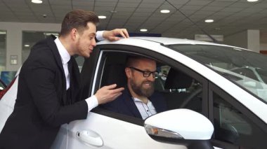 Gösterişli siyah takım elbiseli kendine güvenen bir satış müdürü, galeride şık beyaz bir arabanın içinde oturan olgun bir beyaz adama tutkuyla danışıyor. Etkileyici etkileşim, sergi salonlarıyla birlikte mükemmel aracı seçmenin heyecanını vurgular.