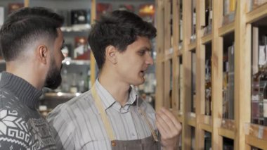 Önlük giymiş odaklanmış bir erkek şarap imalatçısı, sıcak bir şarap dükkanının raflarını araştıran bir müşteriye dikkatle yardım ediyor. Sıcak ortam ve düzgünce düzenlenmiş şişeler şarap meraklıları için mükemmel bir seçim için davetkar bir ortam yaratıyor.