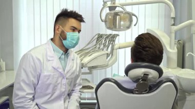 Ağız maskesi ve eldiven takan genç bir beyaz dişçi, modern bir diş muayenehanesinde müşterisinin dişlerini dikkatle inceler. Çekici esmer doktor, bu klinik alanda profesyonellik ve özen gösteren tepsiden ustalıkla bir sonda alıyor.