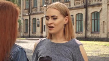 Sarışın bir kadın, elinde kağıt kahve fincanları olan kızıl saçlı arkadaşıyla hareketli bir şekilde sohbet ediyor ve çarpıcı bir üniversite arkaplanının önünde neşe saçıyor. Gülüşleri havayı dolduruyor, hikayelerini paylaşıyorlar, dostluğun ve gençliğin özünü somutlaştırıyorlar.