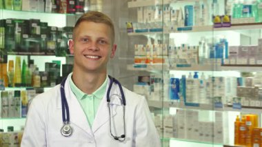 Neşeli bir erkek doktor, elinde bir paket tutarken parlak bir eczanede kameraya gülümsüyor. Dostane tavrı ve profesyonel giyimi, düzgünce düzenlenmiş ilaç ürünlerinin arka planına göre ayarlanmış, davetkar bir atmosfer yaratıyor.