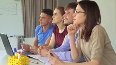 Canlı yaratıcı bir ekip öne doğru eğiliyor, modern ofis ortamındaki bir sunuma hayran kalıyorlar. Bu yakın plan çekimde dört çekici profesyonel kadın ve iki adam renkli kalemler ve bir dizüstü bilgisayarla çevrili şık bir masada oturuyorlar.