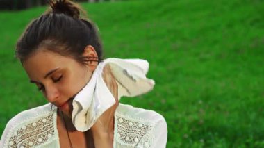 Açık hava yoga seansından sonra, at kuyruklu, koyu renk saçlı genç bir kadın yüzünü terden parlayarak siliyor. Etrafı yemyeşil çimlerle çevrili, dinginliği ve kararlılığı somutlaştırıyor, sağlıklı bir yaşam tarzının güzelliğini gösteriyor.