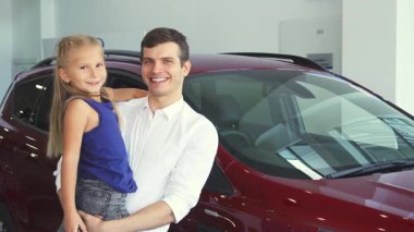 Parlak bir araba galerisinde, neşeli bir baba gülümseyen kızını kollarına alır, her ikisi de mutluluk saçar. Arka plandaki parlak, parlak araba bu iç açıcı aile anına modern bir zarafet katıyor.