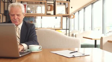 Şık bir kafede, iki iş ortağı güven ve işbirliğini sembolize eden sıkı bir tokalaşmaya girişirler. Genç adam, bakımlı sakallı, kendinden emin bir şekilde yaşlı arkadaşının yanında oturuyor, o da elinde bir kalemle panoya işaret ediyor,