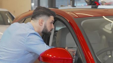 Kendine güvenen, sakallı bir adam galeride çarpıcı bir kırmızı arabayı incelerken kameraya ışıldıyor. Onun şık giyimi ve çekici gülümsemesi yeni otomotiv seçeneklerini keşfetmenin heyecanını yansıtıyor, tüketiciliğin heyecanını ve neşeyi somutlaştırıyor.