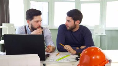 Doğal ışıkla dolu parlak bir ofiste, iki odaklanmış erkek mimar canlı bir tartışmaya girişiyor, planlar ve bir dizüstü bilgisayarın üzerinde hareketli bir şekilde hareket ediyorlar. Esmer, sakallı mimar iş arkadaşlarının kulağını incelikle işaret ediyor.